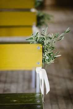 an empty yellow bench with white ribbon tied around it and some green plants in the back