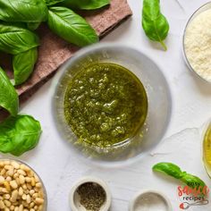 pesto sauce in a bowl surrounded by other ingredients on a white counter top with basil leaves