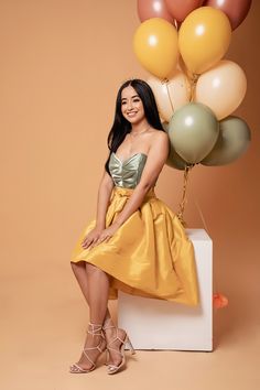 a woman in a yellow dress sitting on a white box with balloons behind her and smiling at the camera