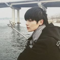 a young man with black hair wearing a coat and scarf standing next to the water