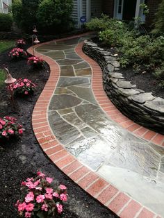 a brick walkway in front of a house with pink flowers on the side and green grass