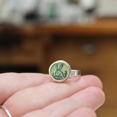 This adorable Rabbit ring has a vintage storybook quality.Available in whole sizes 6 through 9I carved the sterling silver setting from a block of wax, then cast that wax pendant into metal and molded it so I can make multiples. Both sides of the setting then get a layer of vitreous enamel (fine glass) and fired at 1400 degrees in a kiln, four separate times, to build strength and color saturation . Finally I take an illustration from my sketchbook, turn it into an enamel screen print, and fire Rabbit Ring, Vintage Storybook, Vitreous Enamel, Build Strength, Dog Jewelry, Enamel Necklaces, Enamel Bracelet, Enamel Ring, Charm Rings