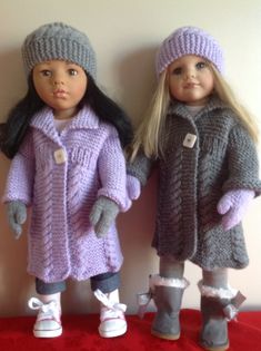 two dolls are standing next to each other on a red blanketed surface, one is wearing a purple coat and the other has a gray hat