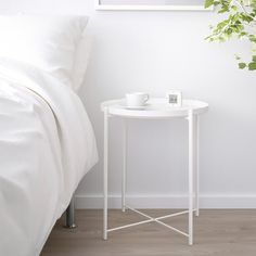 a small white table with two cups on it next to a bed and plant in the corner