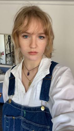 a woman with blue eyes wearing overalls and a white shirt is looking at the camera