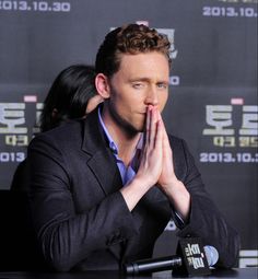 a man sitting at a table with his hands clasped in front of his face and two microphones behind him