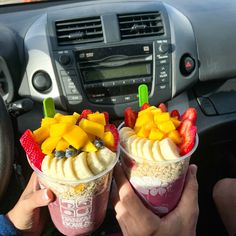 two people holding up cups with fruit and yogurt on them in front of a car