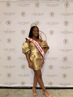 a woman standing in front of a white backdrop wearing a gold dress and holding a red sash