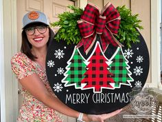 a woman holding up a merry christmas sign in front of a door with a wreath on it