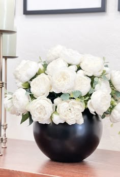 a black vase filled with white flowers on top of a wooden table next to a lamp