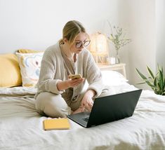 a woman sitting on a bed looking at her cell phone and laptop