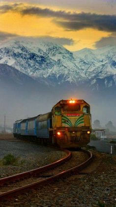 a train is traveling down the tracks in front of snow - capped mountain tops and clouds