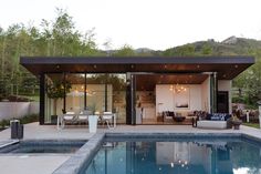 an outdoor living area with a pool and dining room in the back ground, surrounded by greenery