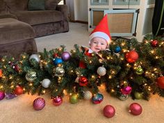 a baby in a christmas hat is peeking out from behind a tree with ornaments on it