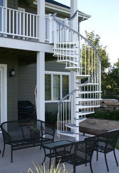 a white spiral staircase in front of a house with black chairs and tables on the patio