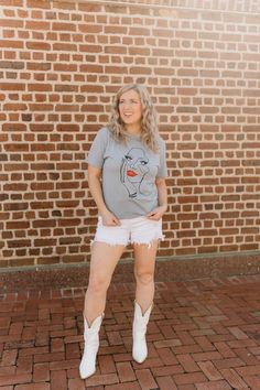 a woman standing in front of a brick wall wearing white shorts and a gray shirt