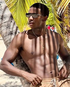 a shirtless man standing next to a palm tree on the beach with his hands in his pockets