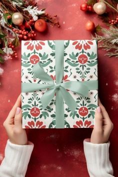 a person holding a wrapped present in front of christmas decorations and greenery on a red background