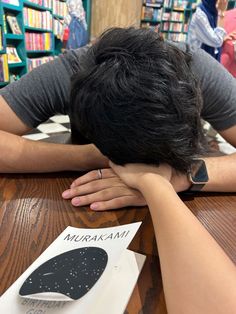 a person with their head on the table in front of bookshelves and people