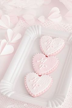 three heart shaped cookies sitting on top of a white plate next to pink flowers and petals