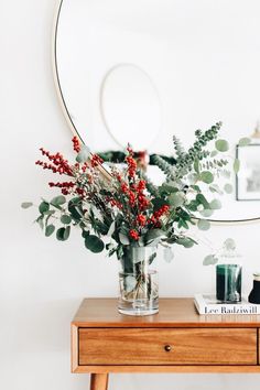 a vase filled with red berries and greenery on top of a wooden table next to a mirror