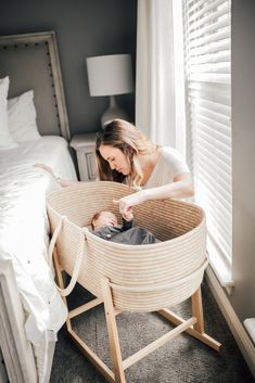 a woman holding a baby in a wicker bassinet next to a bed