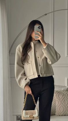 a woman taking a selfie while wearing black pants and a white jacket with a gold chain bag