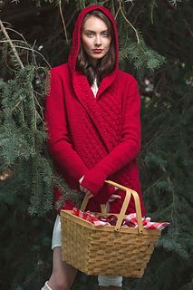 a woman holding a basket full of apples in front of some pine tree branches and wearing a red hooded sweater