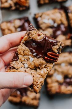 someone holding up a chocolate chip cookie bar with pecans in the foreground and more cookies in the background