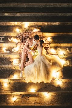 a bride and groom kissing on the steps with fairy lights all around them at night