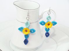 a pair of blue and yellow flower earrings sitting on top of a white plate next to a cup