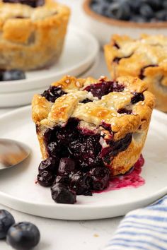 blueberry pie on a white plate with spoons next to it and another pie in the background