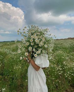 a woman in a white dress holding a bunch of daisies on her shoulder and looking at the camera