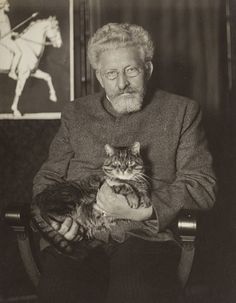 an old man sitting in a chair holding a cat and looking at the camera with a horse behind him