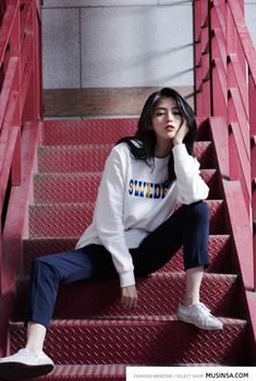 a young woman sitting on the steps in front of red railings, wearing a white sweatshirt and blue pants