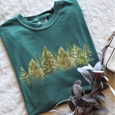 a t - shirt with trees painted on it next to some shoes and a plant