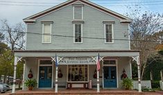 an old white house with blue doors and american flags on the front porch, along with other buildings