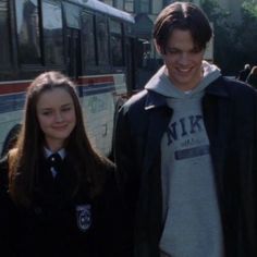 two young people standing next to each other in front of a bus on the street