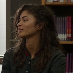 a woman with long hair sitting in front of a bookshelf and looking off to the side