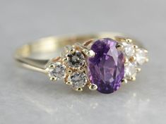 "Beautiful, classic and timeless, this vintage ring is the perfect setting for the pretty, blackberry hued sapphire we've set at the center. Purchased on one of our frequent buying trips to Sri Lanka, or old Ceylon, this purple sapphire has outstanding flash and glitter, perfectly balanced by the tapered diamond shoulders. Metal: 14K Yellow Gold Gem: Purple Sapphire 1.43 Carats Gem Measurements: 5.2 x 7.8 mm, Oval Accents: 6 Diamonds totaling .36 Carats, H in Color, SI in Clarity Ring Size: 7 Ma Classic Multi-stone Sapphire Ring, Elegant Purple Multi-stone Sapphire Ring, Classic Oval Multi-stone Cluster Ring, Classic Purple Diamond Promise Ring, Classic Oval Cluster Ring With Multi-stone, Elegant Cluster Amethyst Ring For Formal Occasions, Classic Purple Birthstone Ring With Center Stone, Elegant Formal Cluster Amethyst Ring, Elegant Purple Cluster Amethyst Ring