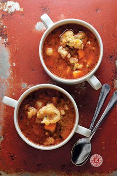 Two cups of vibrant Hungarian cauliflower soup with roasted vegetables, served on a textured red background with silver spoons.