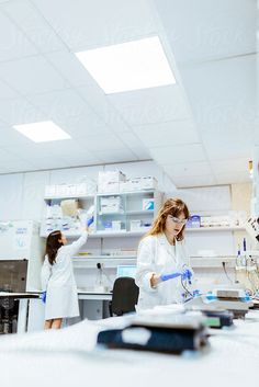 two women in white lab coats working with blue flasks on a table by an open shelf