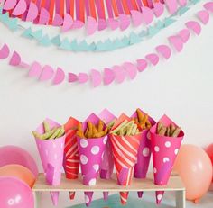 pink paper cups filled with food on top of a wooden table next to balloons and streamers