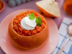 a bowl of chili with sour cream on top sits on a pink plate next to bread and silverware