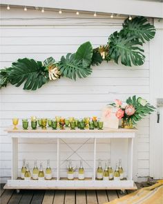 a white table topped with lots of bottles and glasses filled with drinks next to a green plant