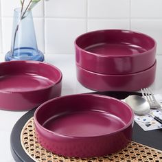 three purple bowls on a table with silverware and flowers in a vase behind them