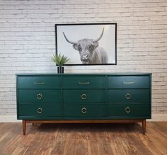 a green dresser sitting in front of a brick wall with a cow painting on it