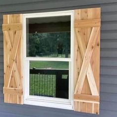 an open window with wooden shutters on the side of a gray house in front of a green yard