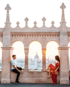 a man and woman sitting on top of a stone wall next to each other looking out at the city