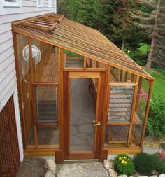 a small wooden greenhouse with glass doors on the front and side of it, next to a house